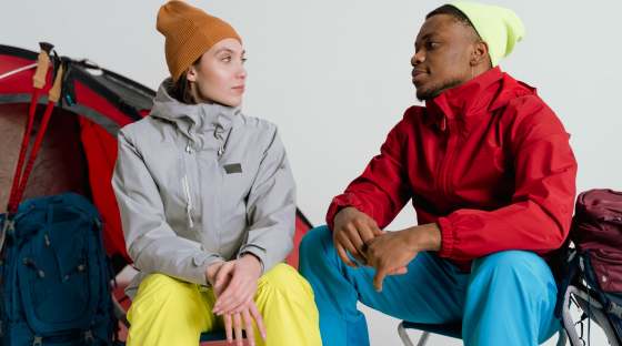 Woman and man sitting in a studio in outdoor clothing with tent behind