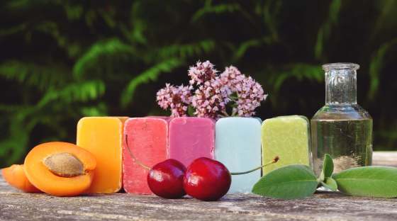 five bars of coloured soap with fruit and herbs next to them