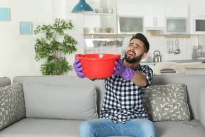 Man sitting on sofa holding bucket with water leak from ceiling