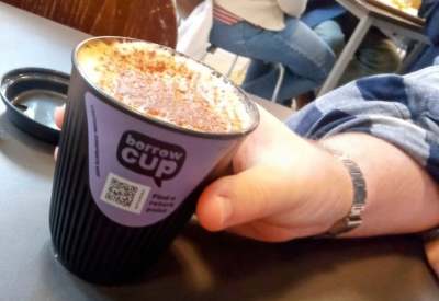 Person's hand and arm holding a Borrow Cup of a hot frothy drink