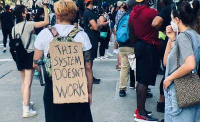 People at a protest with one person with sign on back which reads 'This system doesn't work'