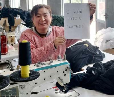 Garment worker at sewing machine surrounded by textiles holding sign which reads 'I make THTC's clothes'