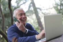 Older man holding mobile phone and using laptop outside