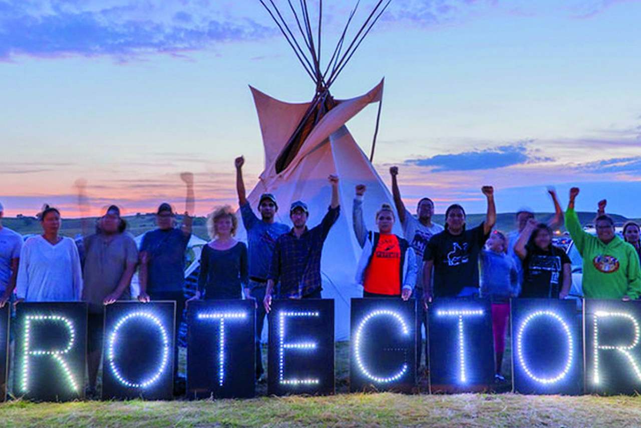 image: indigenous people teepee in background with hands in the air lights spell out the word protectors in foreground