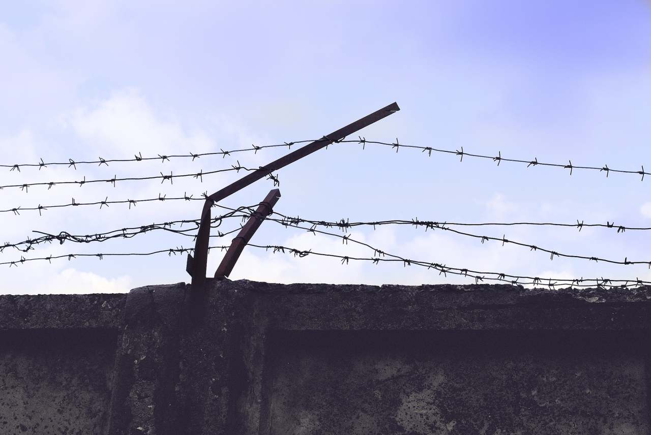 image: barbed wire on a high prison wall in foreground blue sky in background