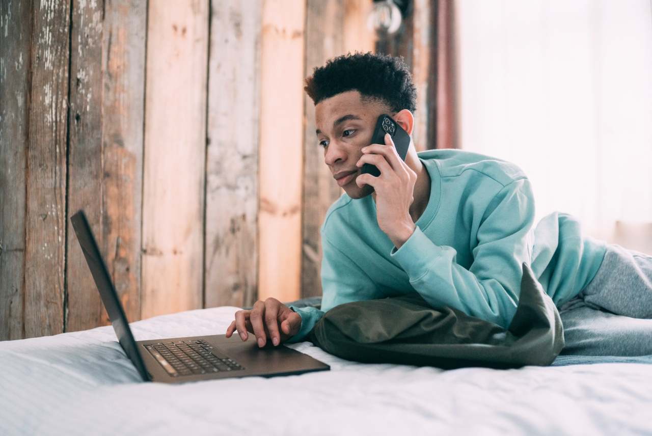 Black man lying on bed with laptop and mobile phone