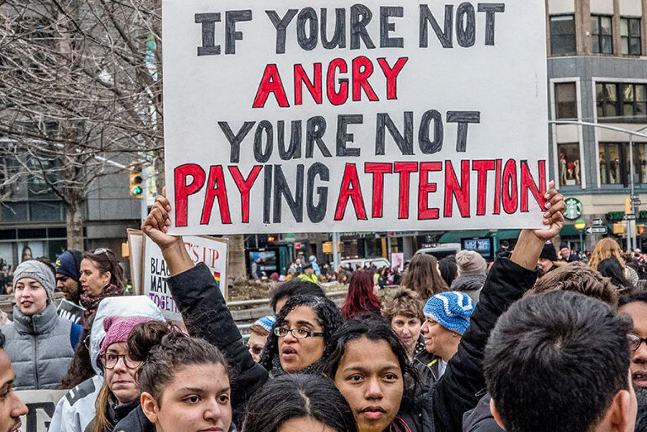 Crowd of people on demo with banner which reads 'If you're not angry you're not paying attention'