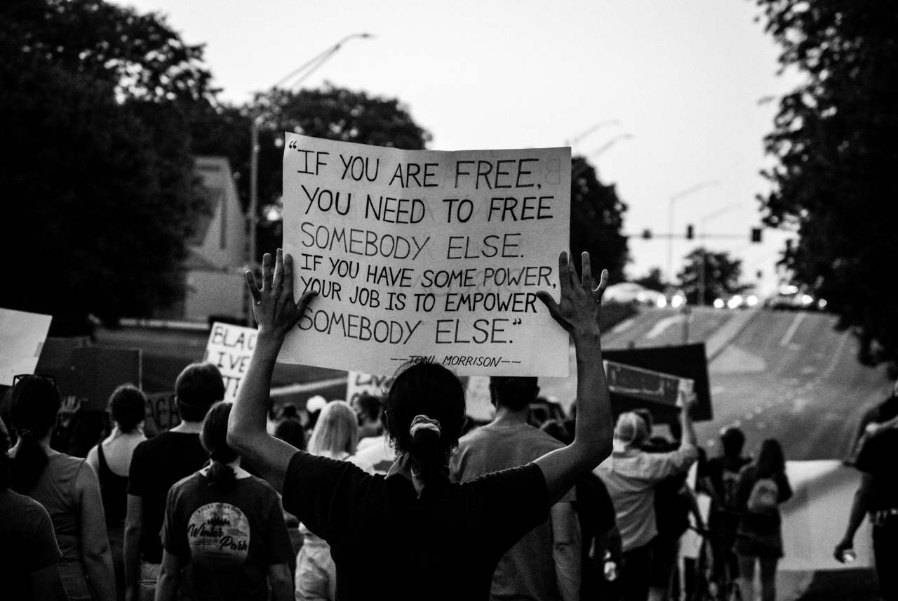 Black and white photo of people protesting. One person's banner reads 'If you are free you need to free somebody else'.