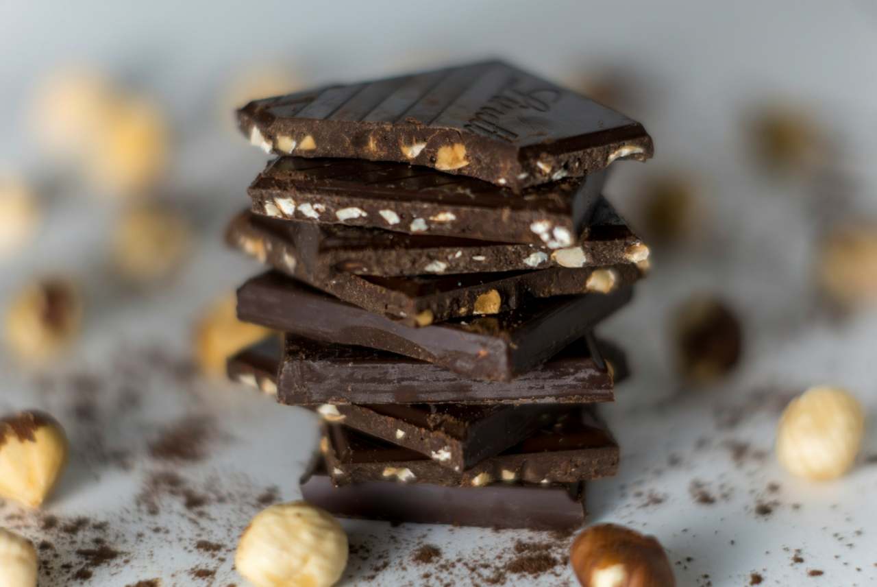 Stack of squares of dark chocolate with nuts scattered around it