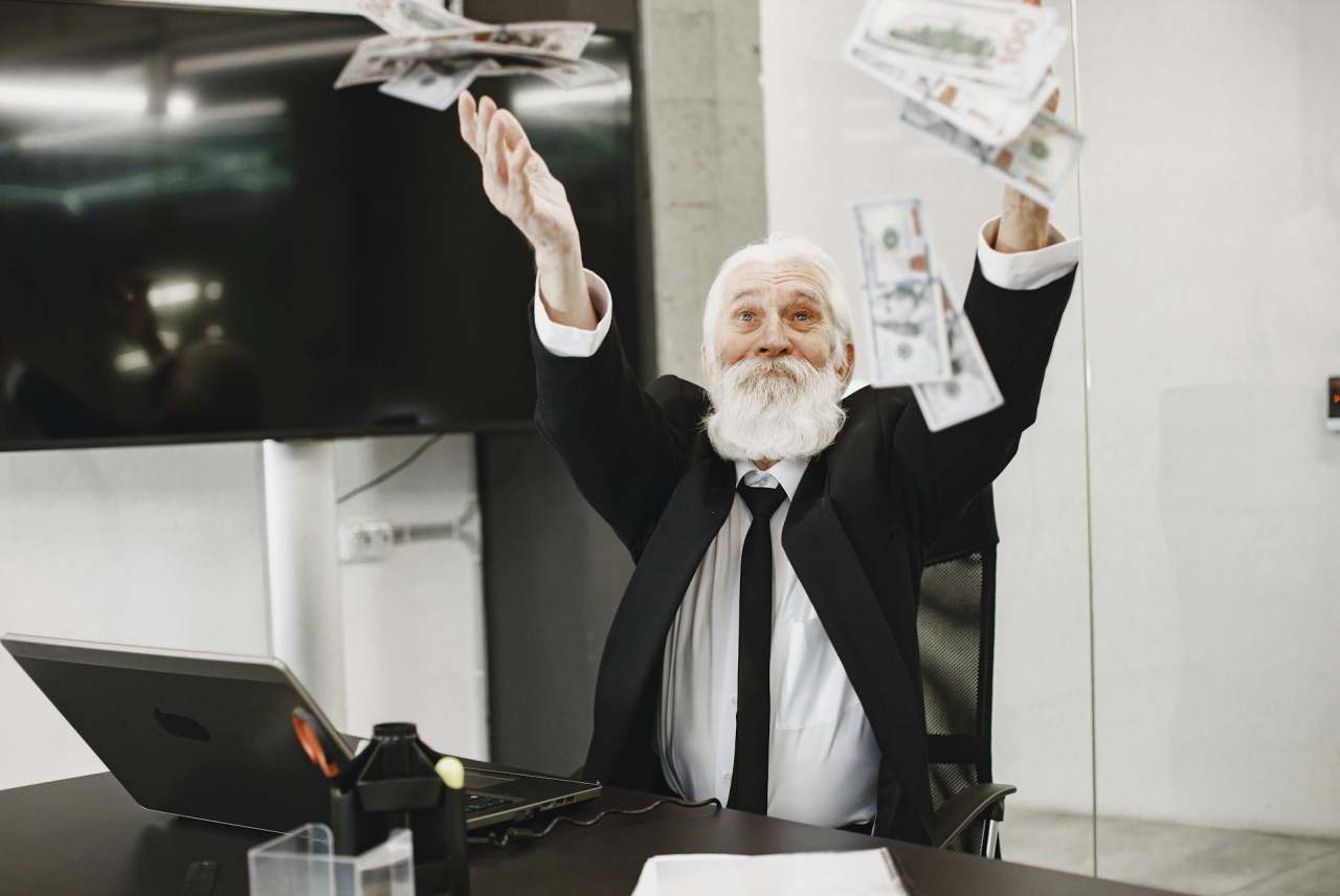 Older white man with beard in office throwing cash notes in air