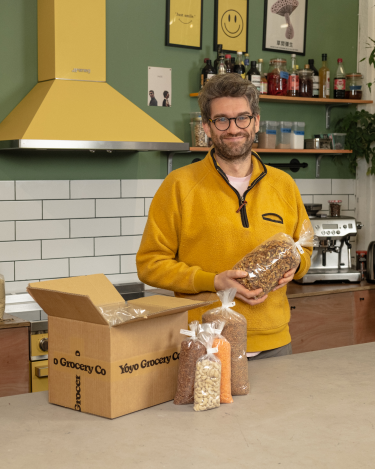Man in yellow top holding packet of food in kitchen with box of other dried food products on counter